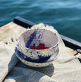 Close up of a hand-painted white and blue bowl on a blanket near a lakeke